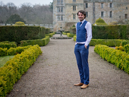 A full body landscape photo of Matt Dovey, dressed in a striped white shirt, blue tie and blue waistcoat, stood on a gravel path with low hedges in with a fountain some distance behind him and a stately home in the background
