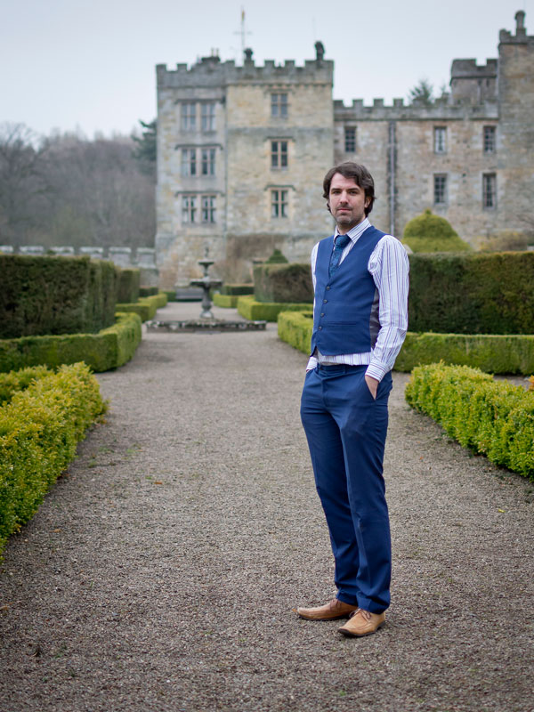 A full body portrait photo of Matt Dovey, dressed in a striped white shirt, blue tie and blue waistcoat, stood on a gravel path with low hedges in with a fountain some distance behind him and a stately home in the background