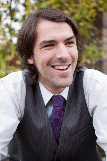 A photo of Matt Dovey, smiling, dressed in a white shirt, black waistcoat and purple tie, photographed against an out-of-focus background of leaves