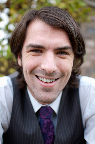 A headshot of Matt Dovey, dressed in a white shirt, purple tie and black waistcoat, looking at the camera and smiling