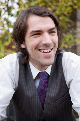 A headshot of Matt Dovey, dressed in a white shirt, purple tie and black waistcoat, looking off to one side and smiling